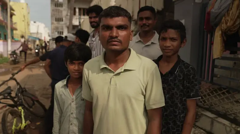 A man stands in a street with children and neighbours gathered behind him