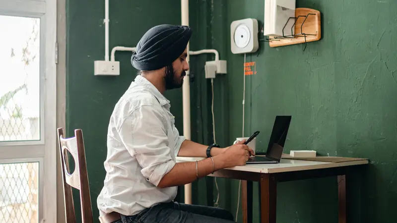 A man in a turban works on a laptop at a small desk against a green wall