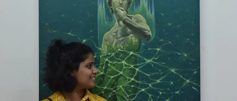 A woman in a yellow shirt smiles beside a large painting of a figure wrapped in green light